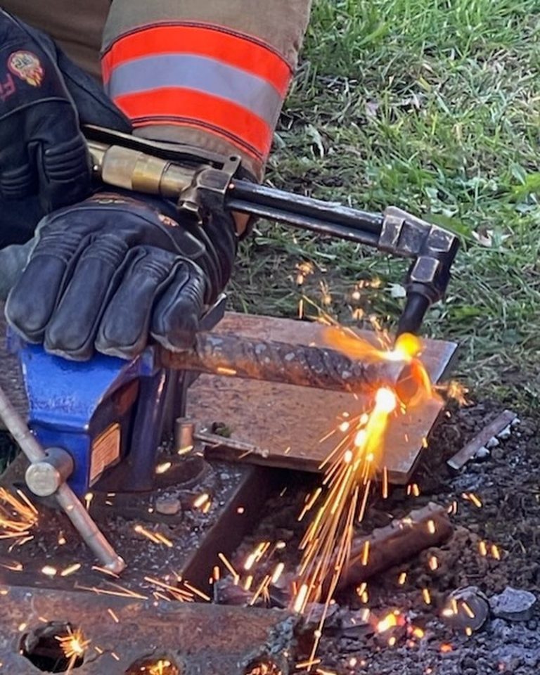 A person wearing gloves uses a welding tool, creating bright sparks as they work on a metal rod held in a blue vise. There is grass in the background.
