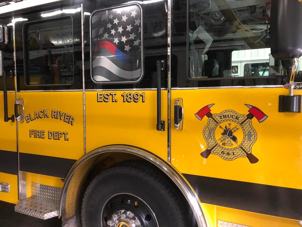 A yellow fire truck with "Black River Fire Dept." and "Est. 1891" on the side. An emblem with crossed axes and the text "Truck 6-6-1" is visible. A flag design is on the window.