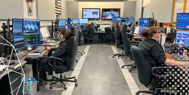 A room with many desks and monitors where people are working. A large announcement reads, "Dispatcher Exam Announced!" with details about filing and exam dates. A website for more information is provided, along with an "Apply Today!" button.