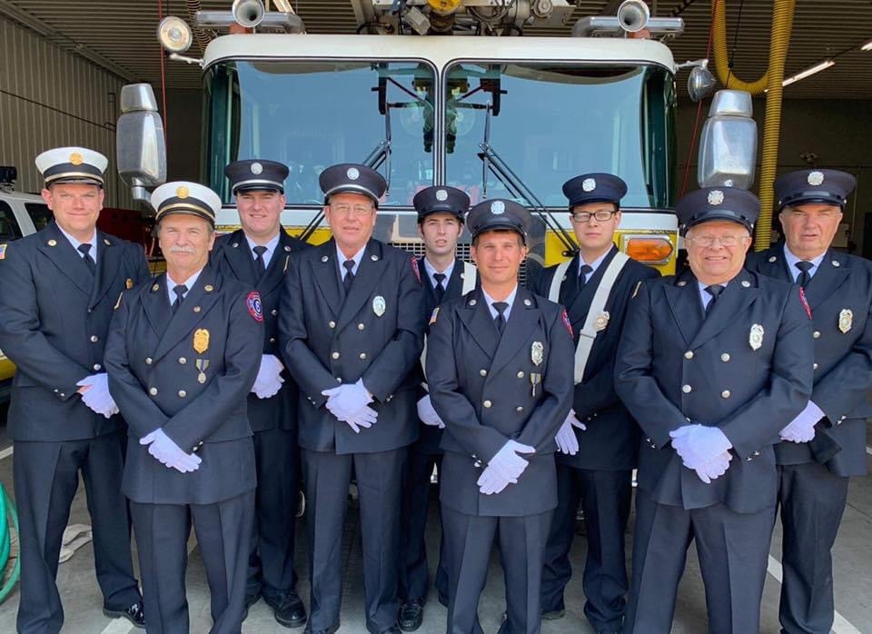 A group of nine firefighters in uniform stand together in front of a fire truck inside a garage. They are dressed formally, wearing matching jackets, caps, and white gloves, and pose in a line facing the camera.
