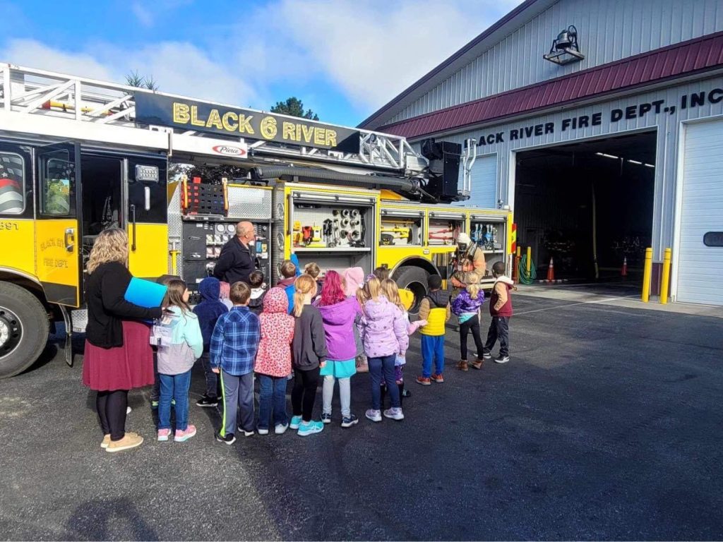 A group of children stands near a yellow fire truck with two firefighters demonstrating its features. They're outside the Black River Fire Dept. building. A woman is present, possibly a teacher or guide. It's a sunny day.