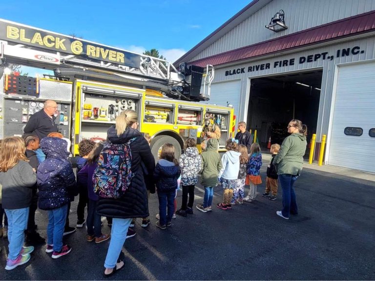 A group of children and a teacher stand in front of a yellow fire truck outside the Black River Fire Department building. A firefighter is showing equipment from the truck. The sky is clear and sunny.