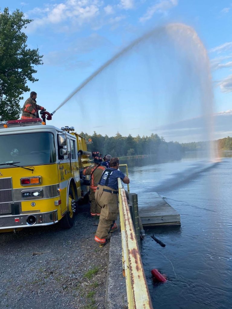 A firefighter on top of a yellow fire truck directs a powerful water stream into a lake. Several firefighters, wearing gear, stand nearby observing. A clear sky and trees are in the background.