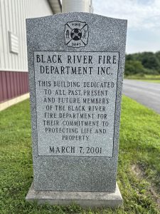 A gray stone monument with an inscription honoring past, present, and future members of the Black River Fire Department for their dedication to protecting life and property. Dated March 7, 2001, with "FIRE DEPT" logo at the top.
