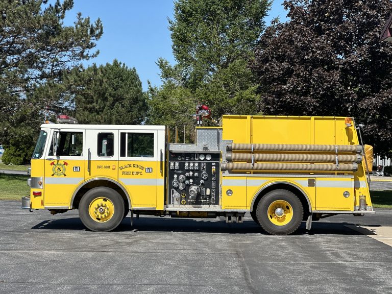 A yellow fire truck parked on a paved area, surrounded by green trees. The truck has multiple compartments and hoses visible on the side. It is a sunny day with shadows on the ground.