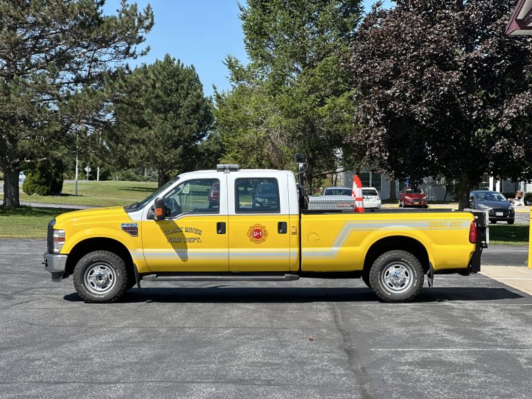 A yellow fire department pickup truck with official insignia is parked on a driveway. The truck has a silver stripe along the side and is surrounded by leafy trees and parked cars.