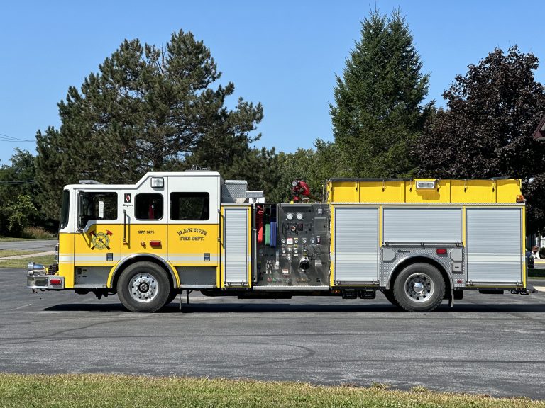 Yellow and white fire truck parked on a paved area, with trees in the background. The truck's side panels are open, revealing equipment and gear stored inside.