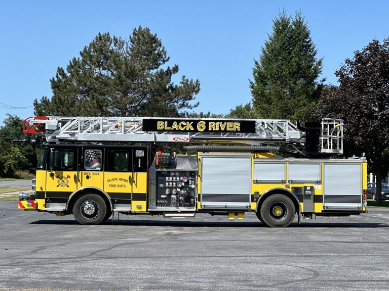 A yellow and black fire truck with "Black River" on the ladder, parked on a paved surface. The truck features various equipment and compartments, with trees and a clear blue sky in the background.