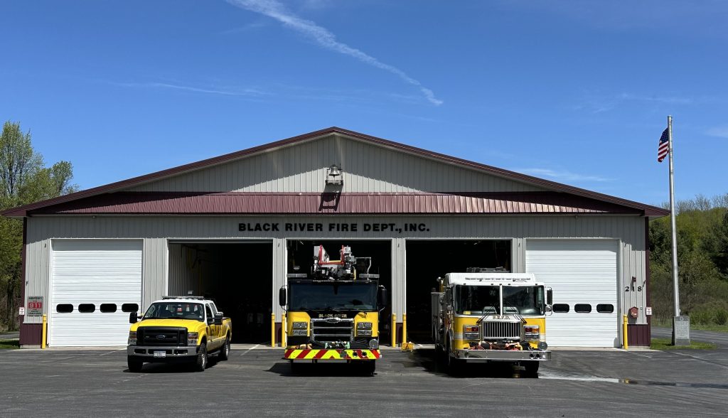Exterior view of Black River Fire Dept., Inc. building with three fire vehicles parked outside: two fire trucks and a yellow pickup truck. An American flag is flying on the right against a clear blue sky.