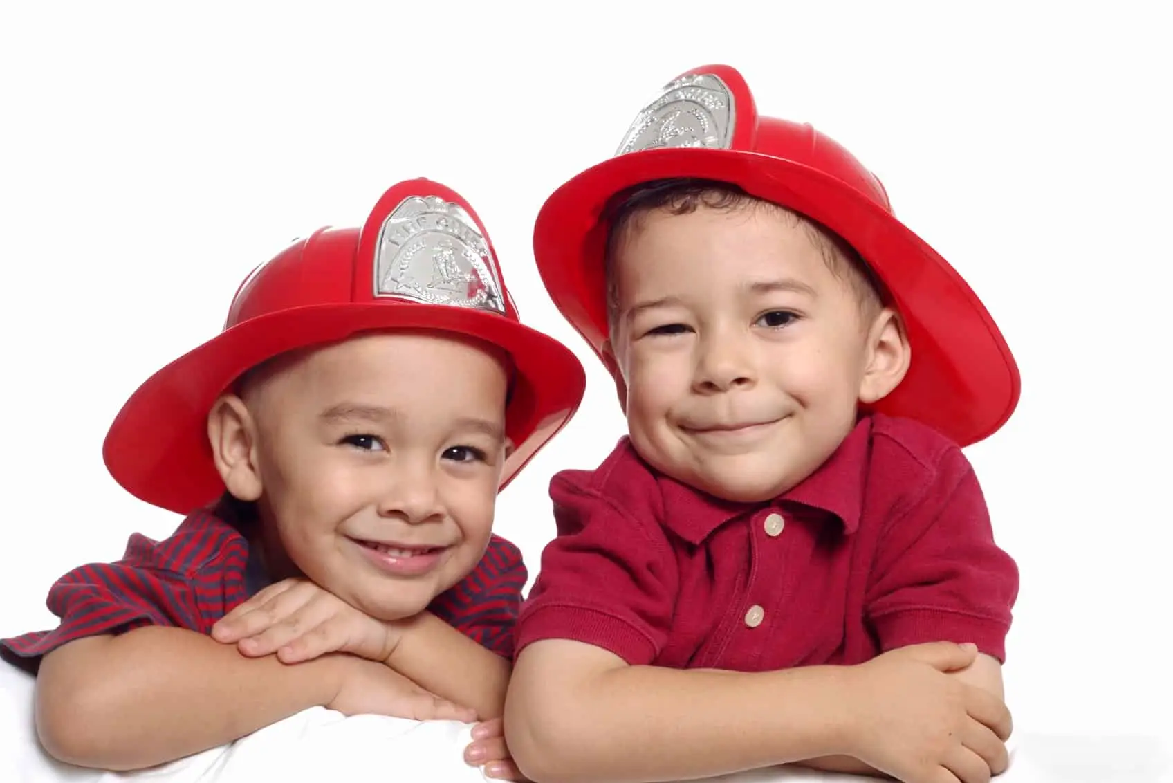 Two young children smiling and leaning on their elbows, wearing red firefighter helmets. They are against a plain white background, dressed in red shirts.