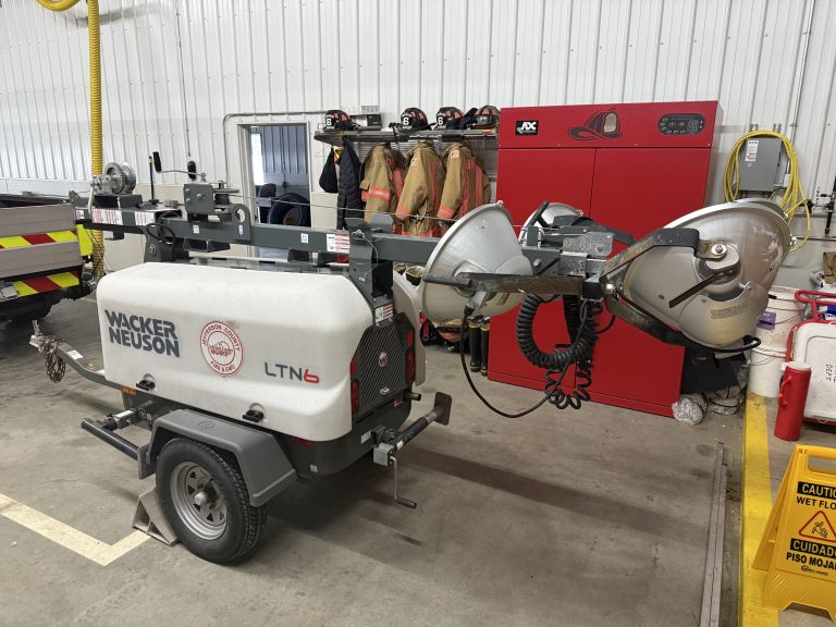 A mobile light tower unit marked "Wacker Neuson LTN6" is parked inside a garage. Firefighting gear hangs in the background, and a red equipment cabinet is nearby.