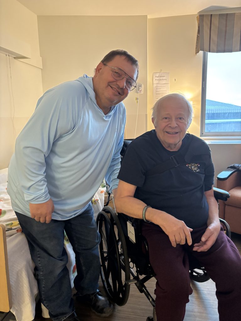 Two men smiling in a room. One man is standing wearing a light blue hoodie, and the other is seated in a wheelchair wearing a dark T-shirt. They appear to be in a healthcare or hospital setting with a bed and window in the background.